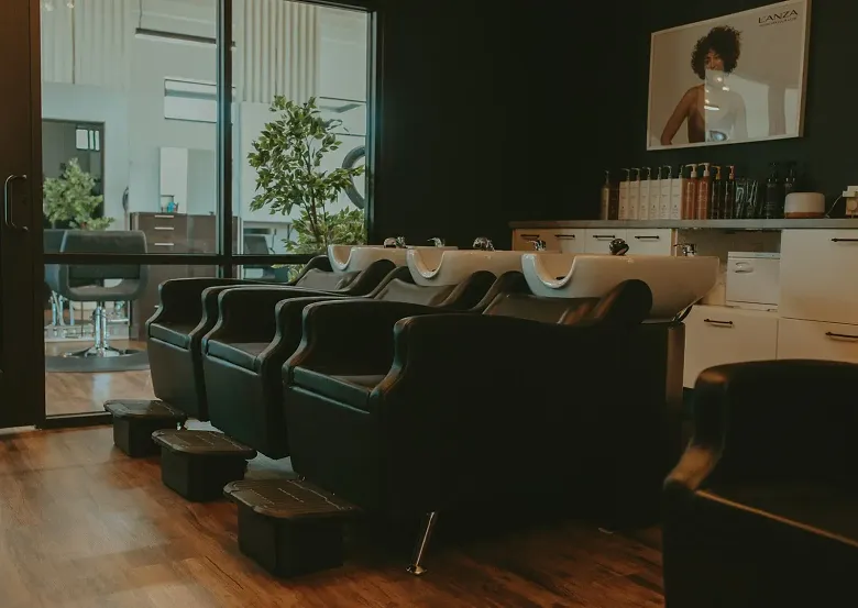 Interior of a hair salon with three black shampoo stations, each with a white sink, black chairs, and black footrests. There are large windows with a view of an outdoor patio area with plants and chairs