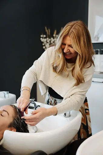 A woman with blonde hair washing another woman's hair at a salon sink